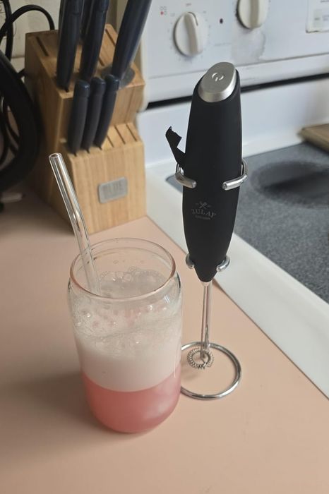 A small kitchen counter setup featuring a frothy pink beverage in a transparent glass, placed next to a handheld milk frother.
