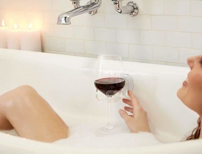 A model soaking in a bubble bath with a clear wineglass holder suctioned to the side of the tub, holding a glass of red wine.