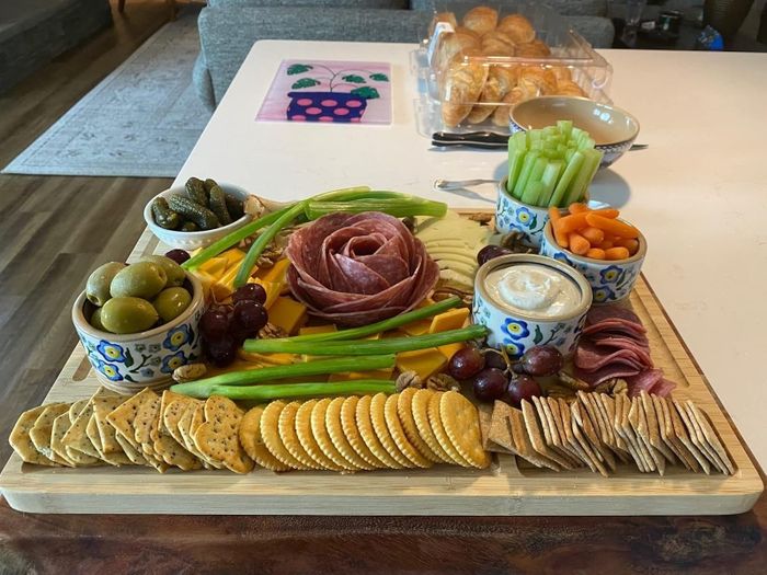 A charcuterie board filled with an assortment of meats, cheeses, crackers, and vegetables arranged in decorative bowls on a wooden surface. Rolls can be seen in the background.