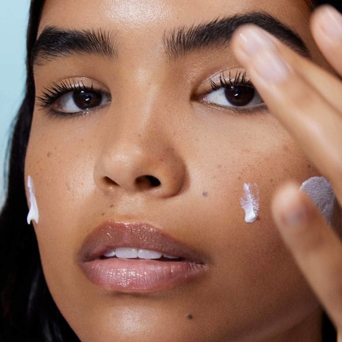 A model applies sunscreen to their face, focusing on the cheek and forehead areas.