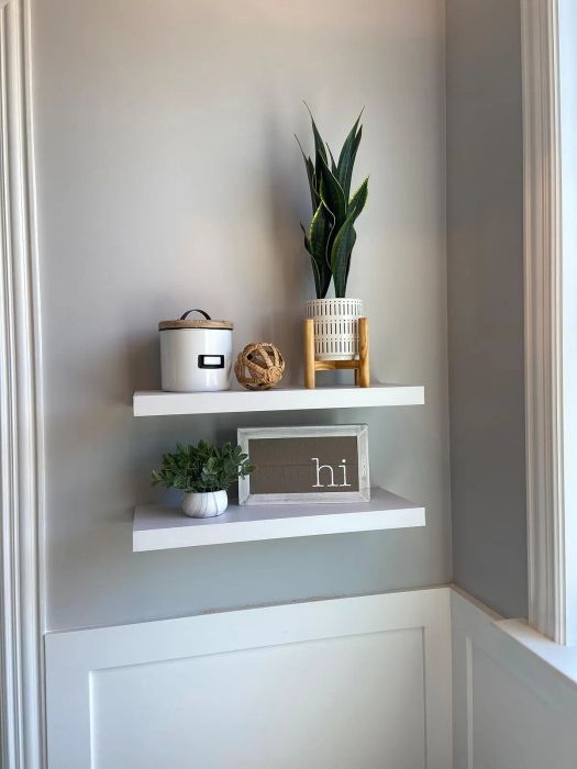 Two white floating shelves holding decorative items: a woven pot with a plant, a white canister, a twine ball, a small plant, and a sign reading