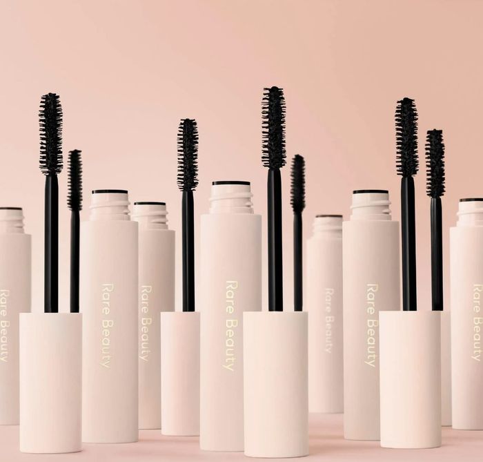 Several tubes of Rare Beauty mascara with wands standing upright on a neutral-colored background