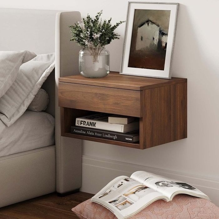 A minimalist nightstand featuring a drawer and shelf, holding books, a plant, and framed art, with an open magazine resting on the nearby bed. The decor reflects a simple yet elegant style.