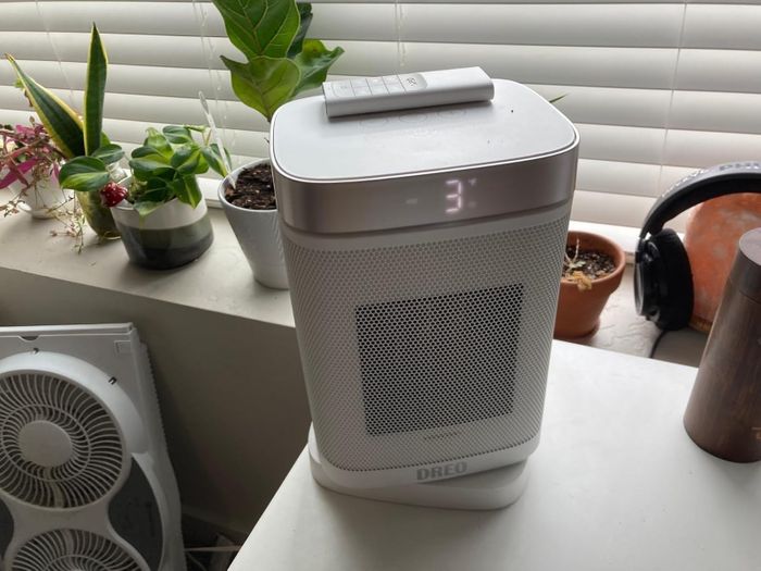 A portable heater placed on a table next to potted plants and headphones, showing the temperature setting.