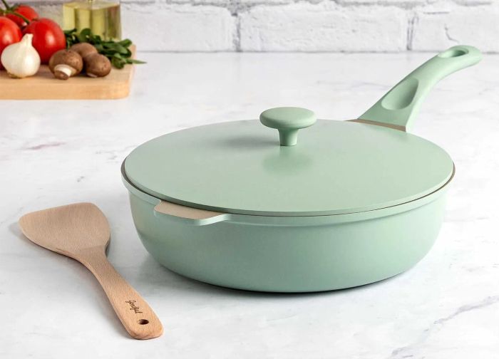 A non-stick sauté pan with a lid and a wooden spoon resting on a kitchen counter with fresh vegetables in the background