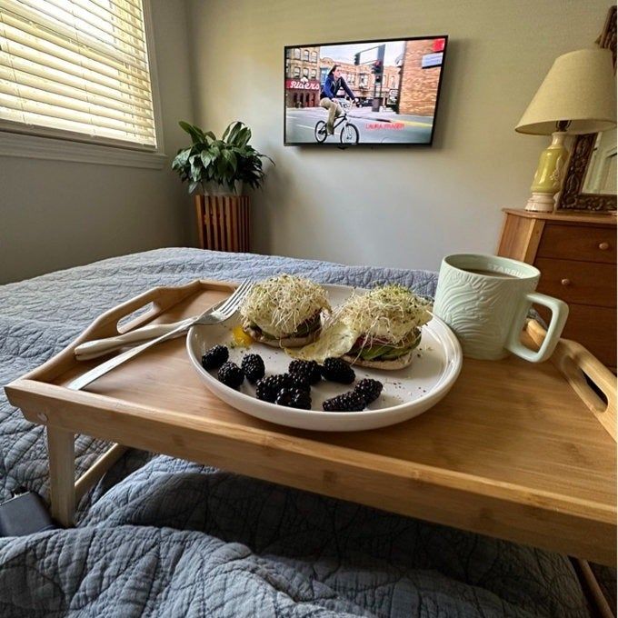 A breakfast tray featuring avocado toast and blackberries placed on a bed, with a TV in the background showing a person cycling. A mug rests on the tray, creating a cozy home atmosphere.