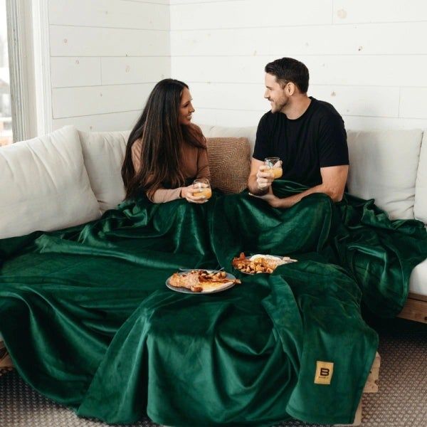 Two people sitting on a couch, enjoying drinks and a charcuterie board, wrapped up in a large blanket. The cozy vibe invites total relaxation.