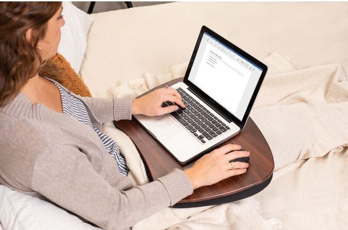 A person working on a laptop while seated on a bed, using a lap desk, creating an ideal environment for both comfort and productivity, perfect for remote work or online shopping.