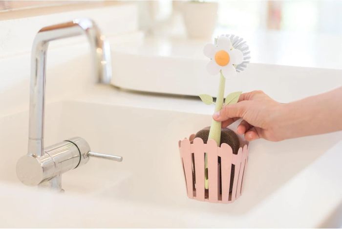A hand places a flower-shaped cleaning brush into a kitchen sink caddy