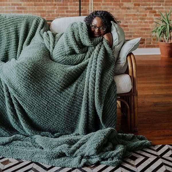 A person cozily wrapped in a large textured blanket on a couch, evoking a sense of comfort and relaxation. An indoor plant and a brick wall provide a warm, cozy backdrop.