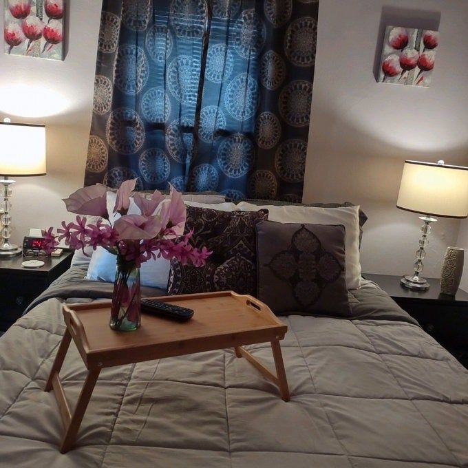 A cozy bedroom scene showcasing a perfectly made bed with decorative pillows, a wooden tray adorned with flowers, and warm ambient lighting from table lamps, creating a tranquil environment.