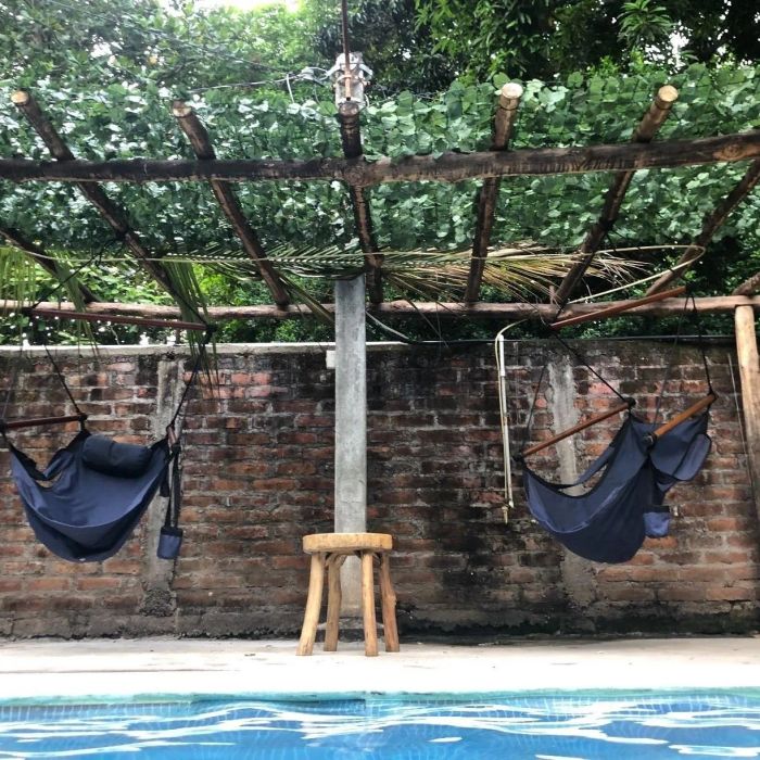Two empty hammocks placed beside a pool, with a rustic wooden stool nestled under a bamboo canopy.
