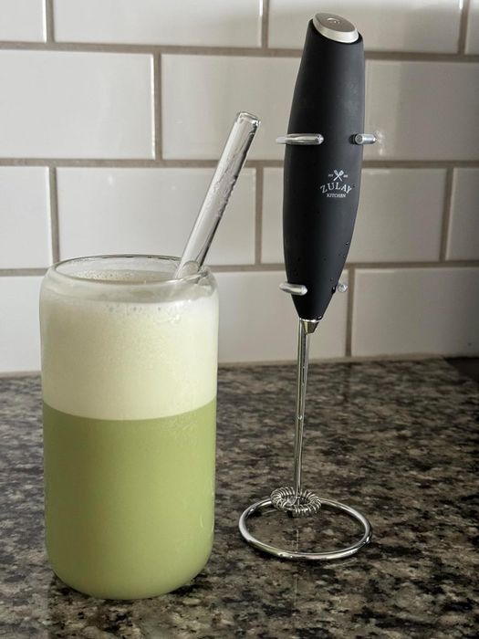 A reviewer's black handheld milk frother placed next to a glass of frothed matcha latte