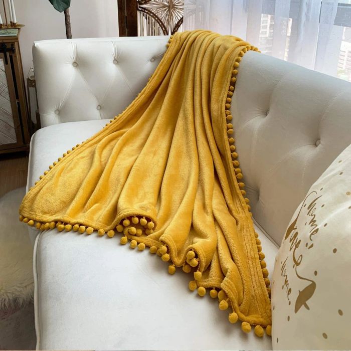 A mustard yellow throw blanket adorned with pom-poms along the edges, casually draped over a couch.