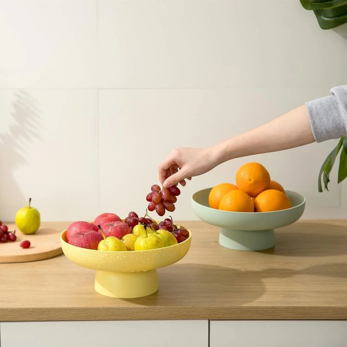 A hand reaches for grapes in a bowl filled with apples and pears, while a bowl of oranges sits in the background on a modern kitchen counter.