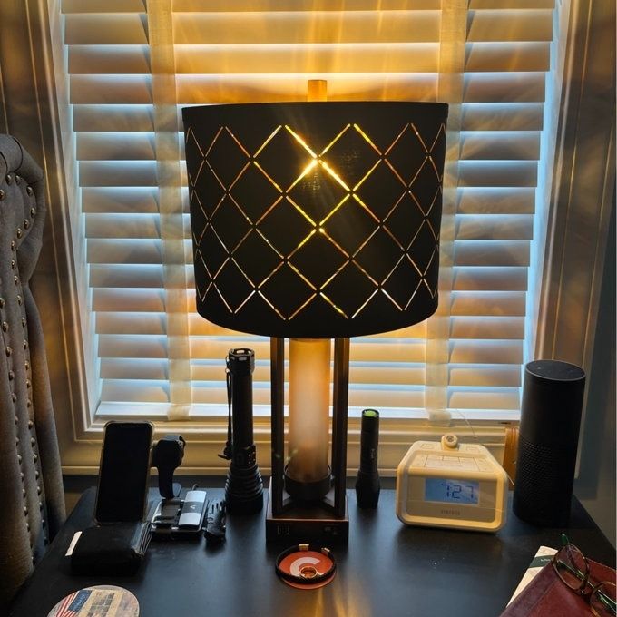 A modern table lamp with a patterned shade, placed on a desk surrounded by gadgets, with window blinds visible in the background. The setting exudes a contemporary, functional ambiance.