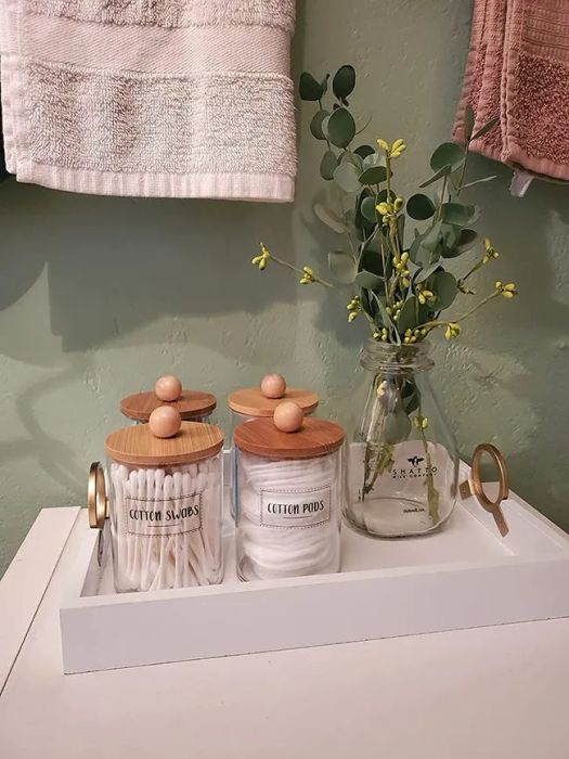 A photo from a reviewer showing labeled glass jars with bamboo lids filled with cotton pads and q-tips, arranged on a tray with a vase of eucalyptus.