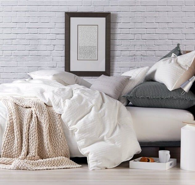 A cozy bedroom featuring a well-arranged bed, layered textured blankets, and pillows set against a white brick wall, with framed artwork hung above, creating a relaxed and inviting atmosphere.