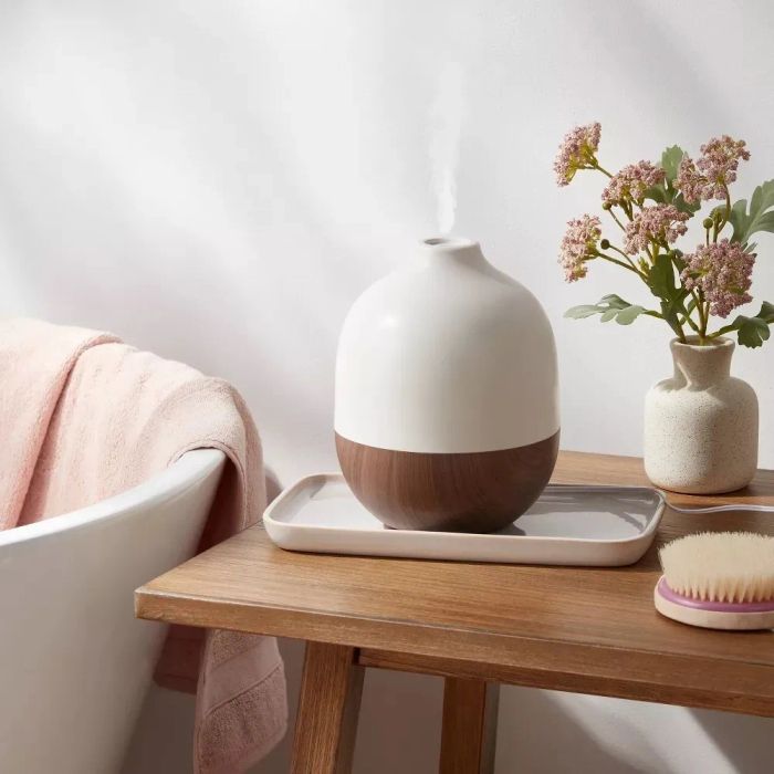 An ultrasonic diffuser placed on a wooden stool next to a flower vase and a brush, releasing a gentle mist for a tranquil atmosphere.
