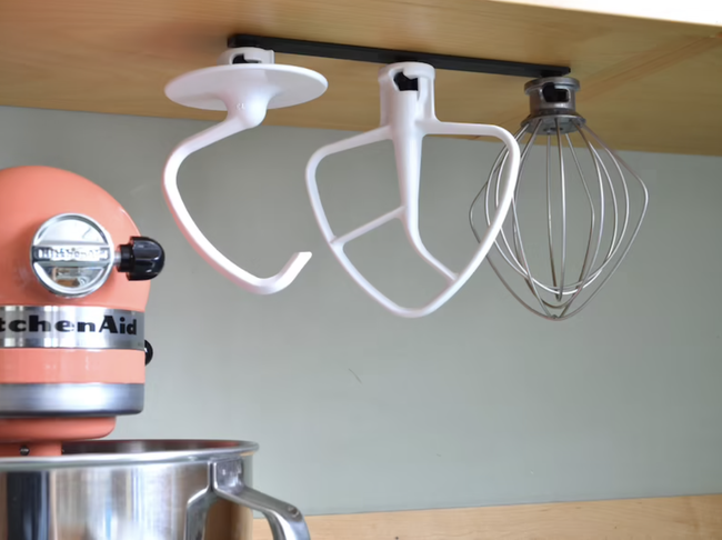 Three KitchenAid attachments suspended from a cabinet by a slim black mount, hanging upside down