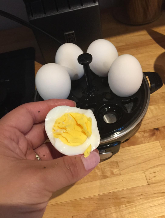 A reviewer demonstrates the inside of a perfectly cooked hard-boiled egg with the black rapid cooker visible in the background.