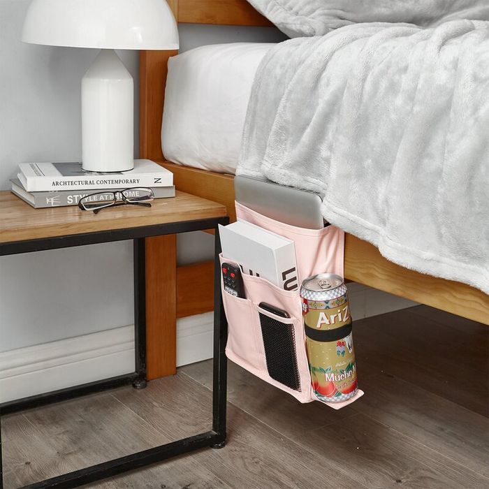 A bedside caddy attached to a bed, storing a can, remote, tablet, and a magazine, placed next to a table adorned with a lamp and an architectural magazine.