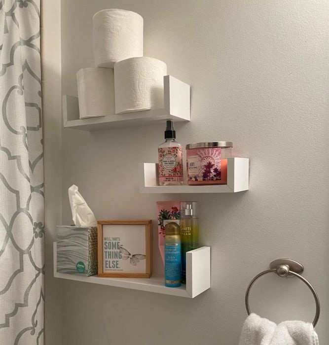 A closeup of a reviewer's three white floating shelves arranged on a bathroom wall, holding toilet paper, tissues, candles, and other bathroom essentials.