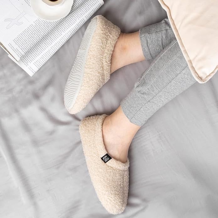 A pair of fuzzy cream-colored sock-style slippers worn by a model.