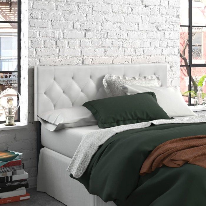 A bedroom featuring a tufted headboard, a white brick wall, a neatly made bed with dark-toned bedding, stacked books, and decorative light bulbs placed on the window sill
