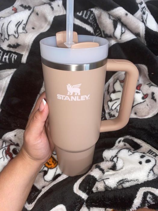 A hand holding a large beige Stanley tumbler with a straw, set against a patterned blanket background.
