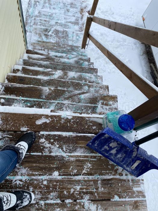 A person standing at the top of snowy wooden steps holding a blue snow shovel, with ice melt visible nearby
