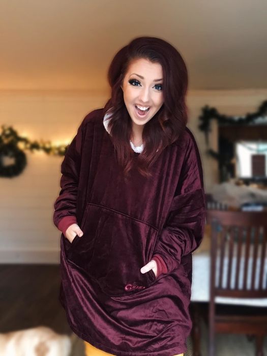 A reviewer models the red hoodie blanket, showcasing its cozy design with a convenient front pouch pocket for extra warmth or storage.
