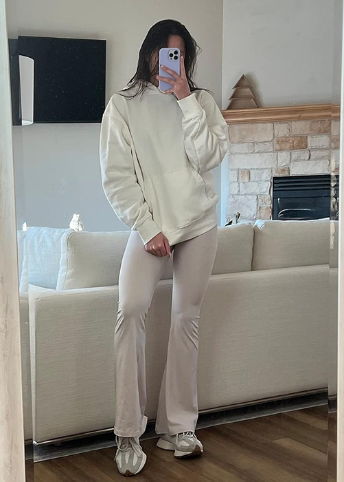 A person captures a mirror selfie while wearing a comfy hoodie and flared pants. They're in a cozy living room, adding to the relaxed vibe.