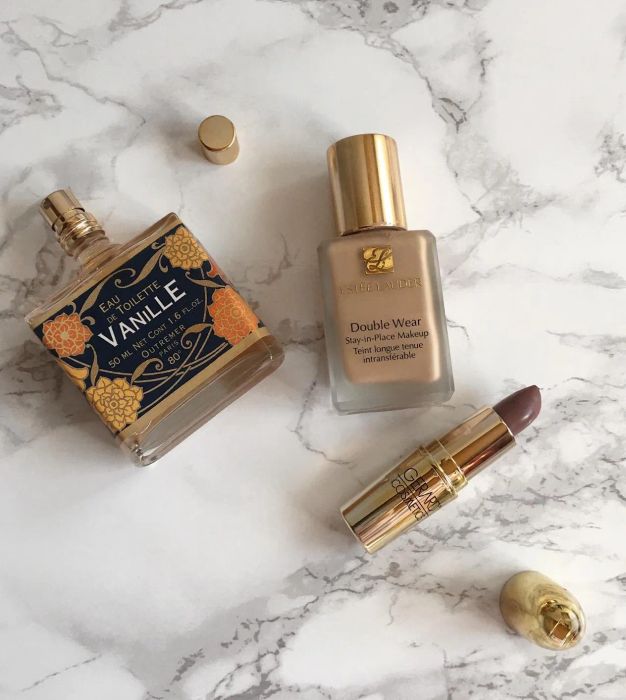Perfume, foundation, and lipstick arranged on a marble surface. The featured items are Eau de Vanille perfume, Estée Lauder Double Wear foundation, and a lipstick.
