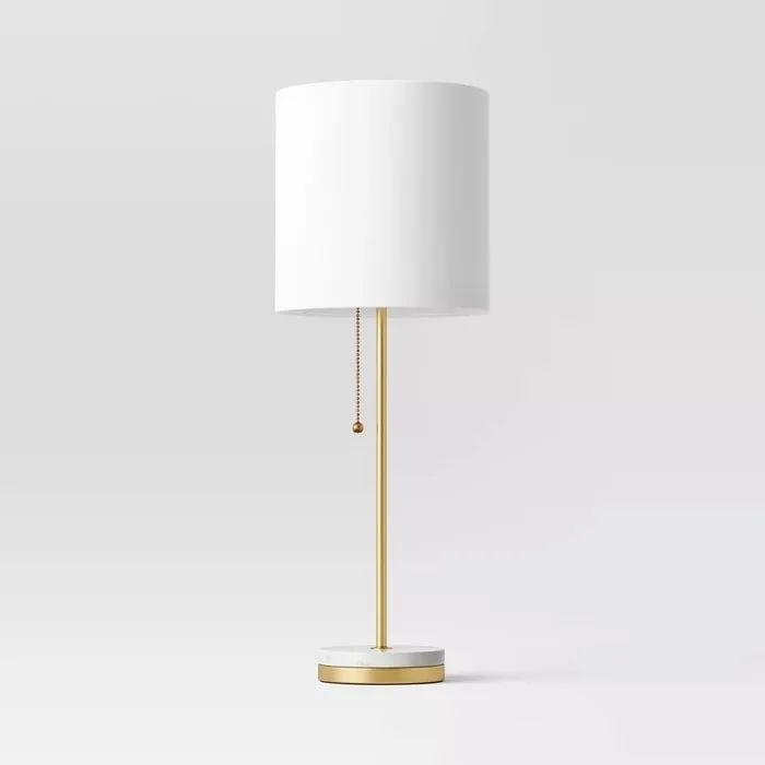 A floor lamp with a white shade and gold base featuring a pull chain, set against a neutral backdrop