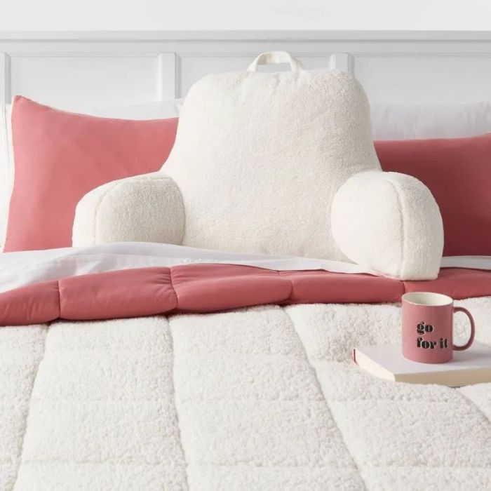 A fluffy backrest pillow positioned on a bed, surrounded by pink pillows, a comforter, and a ‘go for it’ mug. A dream setup for a cozy reading nook.