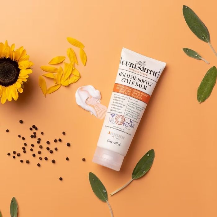 Curlsmith Hold Me Softly Style Balm tube against a peach backdrop, surrounded by a sunflower, seeds, and leaves.