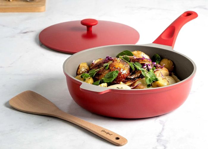 A red non-stick frying pan with a lid, filled with sautéed vegetables, paired with a wooden spatula