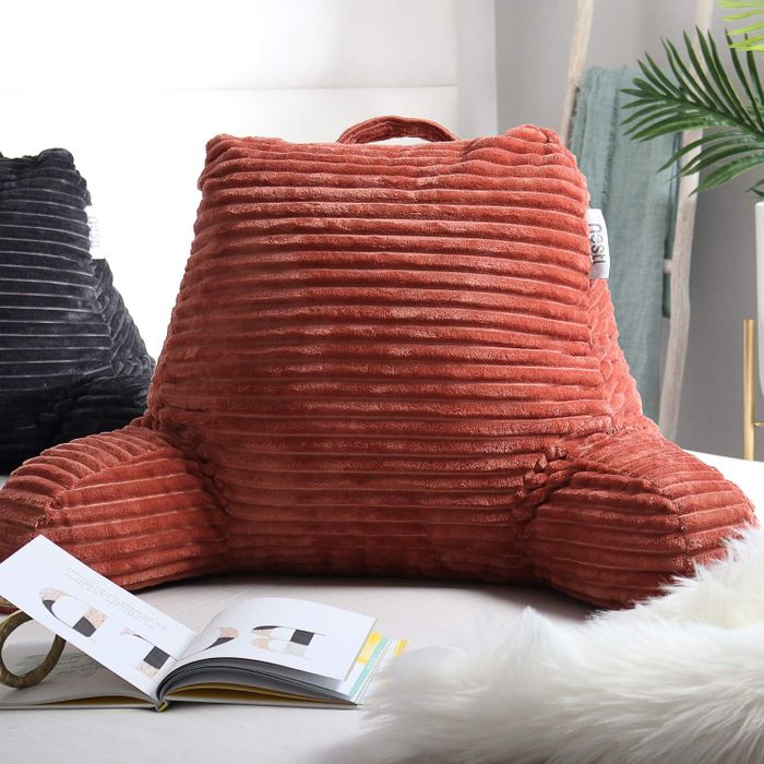 A ribbed backrest pillow with armrests placed on a bed, next to an open magazine and a fluffy rug — perfect for reading or unwinding.