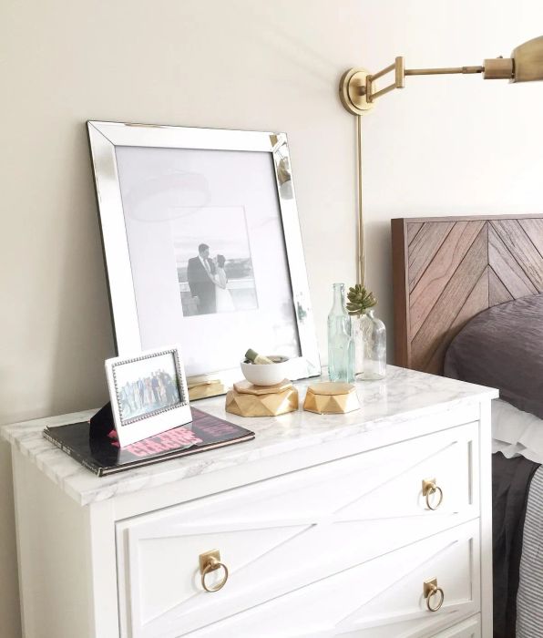 A chic bedroom design featuring a white dresser with gold details, framed photos, decorative bottles, a small potted plant, and a sleek brass wall lamp. The bed is complemented with a patterned headboard and soft pastel pillows.