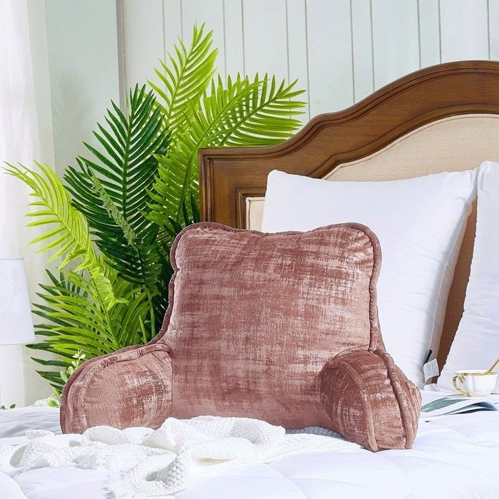 A plush backrest pillow with armrests resting on a neatly made bed, surrounded by pillows and plants, perfect for lounging or reading