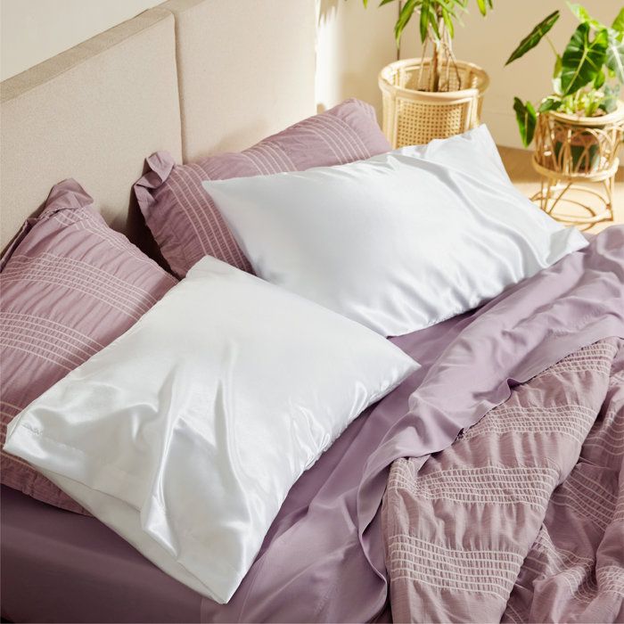 Two white satin pillowcases resting on a bed with textured bedding, accompanied by plants in the background, creating a serene and inviting bedroom atmosphere.