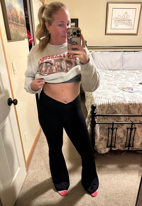 A person taking a mirror selfie in a relaxed outfit featuring a cropped sweatshirt and flared pants, posing in a bedroom with a bed and artwork adorning the walls.
