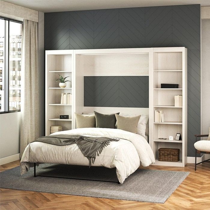 A modern bedroom featuring a Murphy bed, accompanied by shelves. The room is completed with neutral bedding, a cozy throw blanket, and decorative pillows, all set against a chevron-patterned wall.