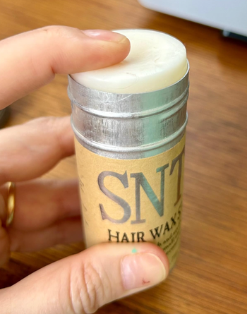 A person holding a container of SNT Hair Wax with the lid partially open.