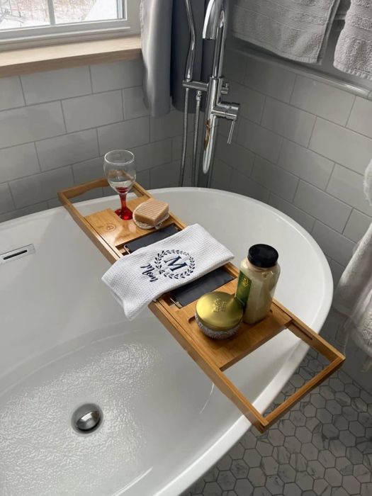 An elegantly organized bathroom featuring a bathtub tray with a candle, soap, loofah, a glass of both red and white wine, and a bottle of bath salts.