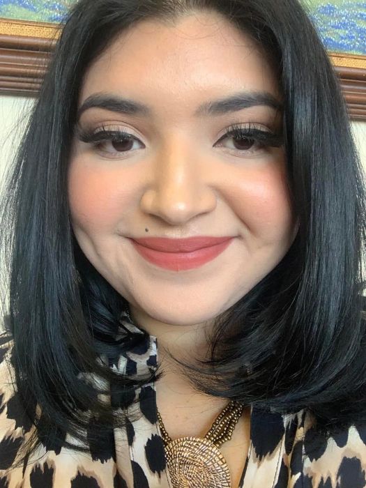 A person smiling, wearing a patterned top and a textured pendant necklace. This image appears in a shopping article