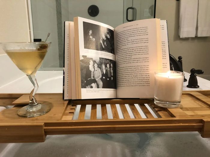 A martini glass, an open book, and a lit candle arranged on a wooden tray over a luxurious bath.