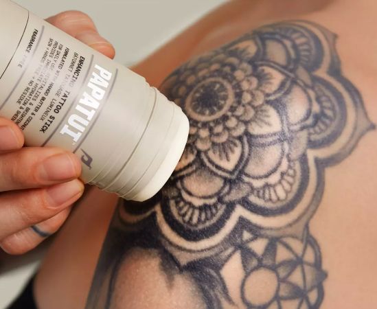 A hand applying Papatu deodorant to a shoulder adorned with a large, intricate black tattoo