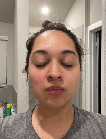 A person with their eyes closed, hair in a bun, wearing a gray shirt, standing in a bathroom, enjoying a moment of relaxation.